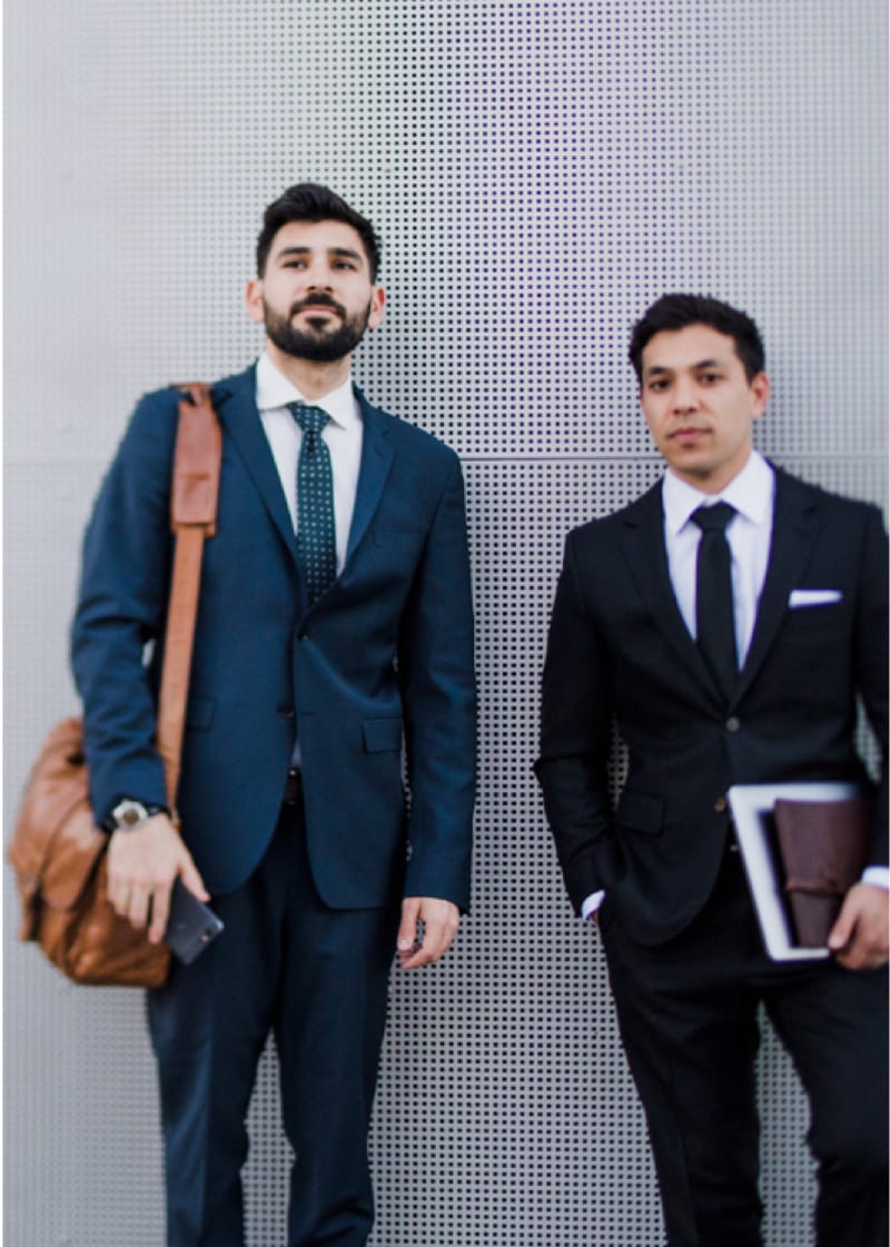 Two nicely dressed service techs standing together Two businessmen posed in front of a wall, wearing nice suits and carrying messenger bags.