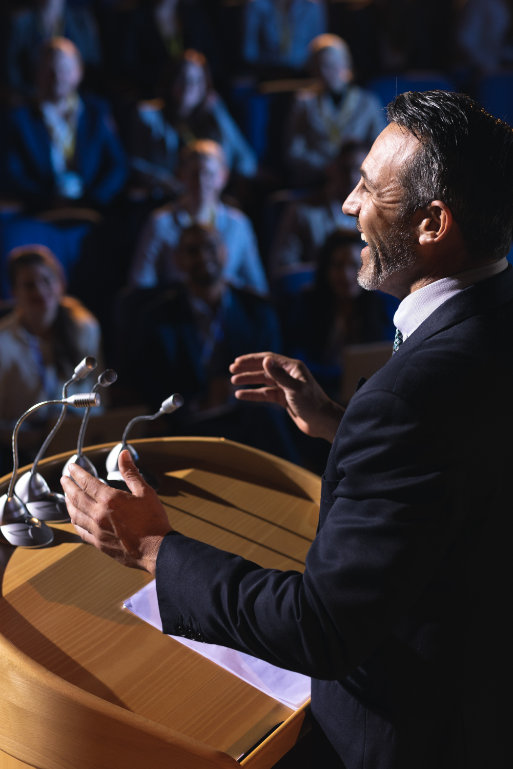 Closeup from the side of a gentleman giving a public speech. He's standing at a podium in front of an auditorium of people.
