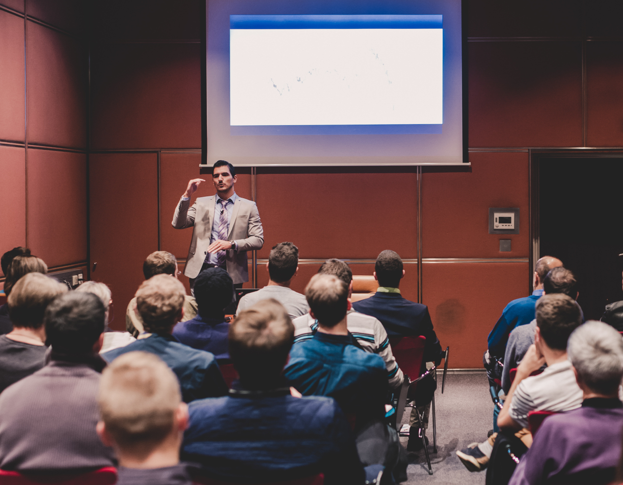 View from the back of a conference room. A man is up on stage at the front, in the middle of speaking to a sea of people that are listening intently to the speaker.